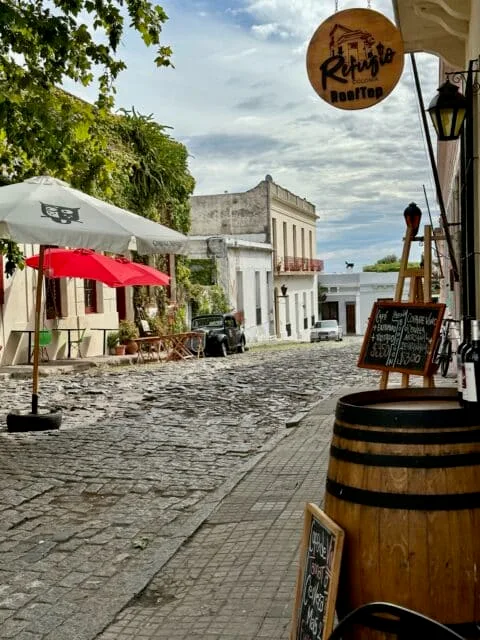 3-day itinerary in Southwest Uruguay. Cobbled street with an antique Ford car and street cafe with outdoor seating and umbrellas for shade. There is also a menu for coffee and wine. Some buildings have iron balconies typical of Uruguay.