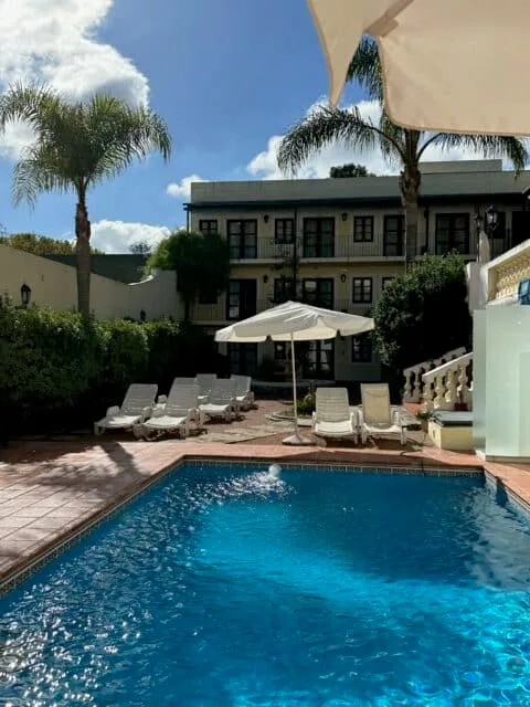 Swimming pool of a hotel with sun loungers and umbrellas for shade. There are two palm trees in a courtyard, in front of the hotel building.