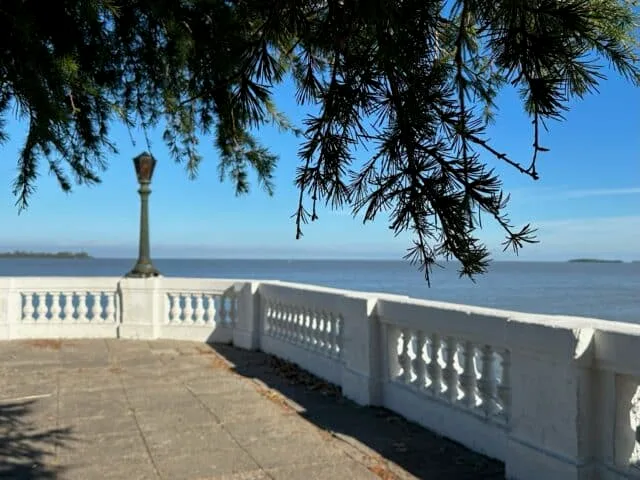 A promenade that has a white stone wall, a lamppost, and trees that overhang above. The blue waters of the river are beyond the wall.