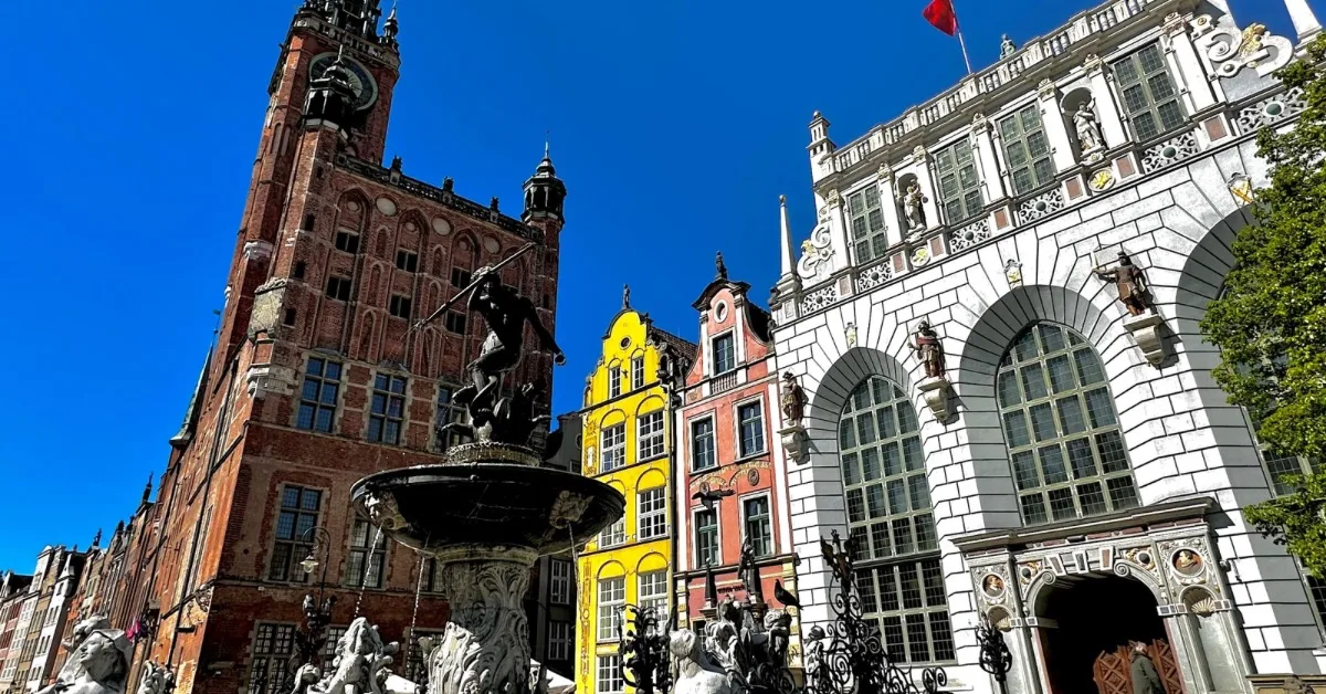 A fountain with Neptune figure on top in front of a red brick church and next to this yellow. Orange and window baroque style buildings in Gdansk.