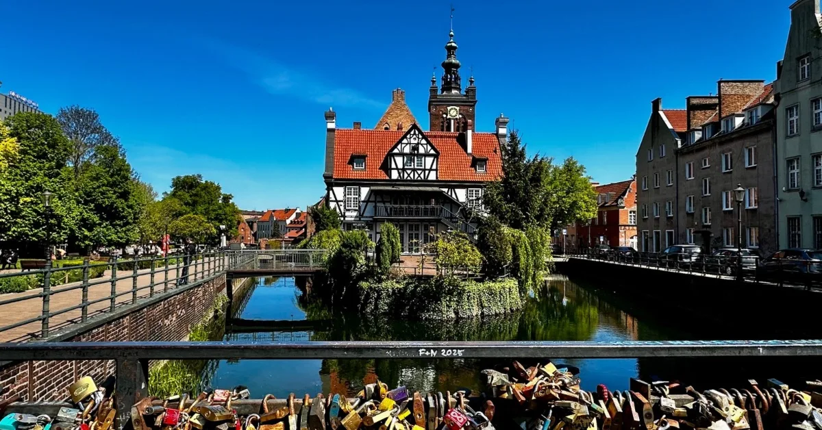 A bridge with multiple locks on it over a canal and a red-roofed building sits on a small island. Behind is a church steeple