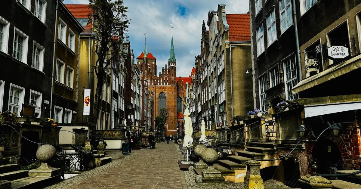 Narrow cobbled street with built up entrances, wrought iron rails and gargoyle headed drains.  At the end of the street is St Mary’s Church built from red brick and with a high green steeple.