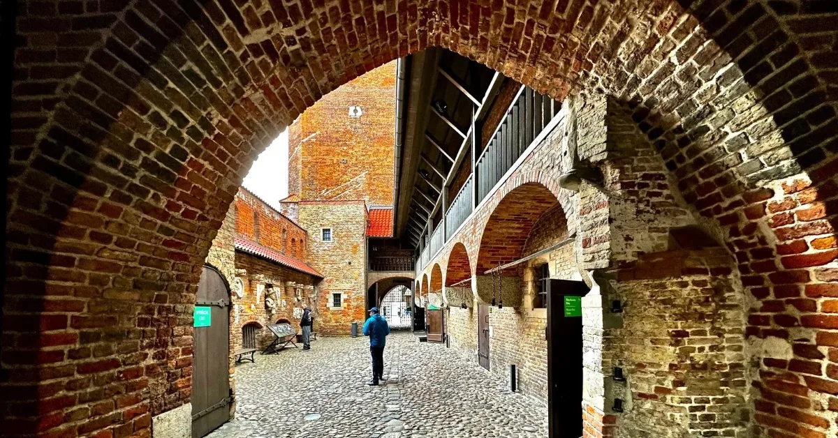 Brick archway leads to a cobbled stone area with a row of arched wall and in front a higher brick tower.