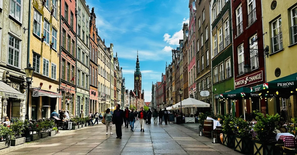 A paved street lined with cafes and outdoor tables and chairs.  Tall rows of buildings each a different colour are on either side.