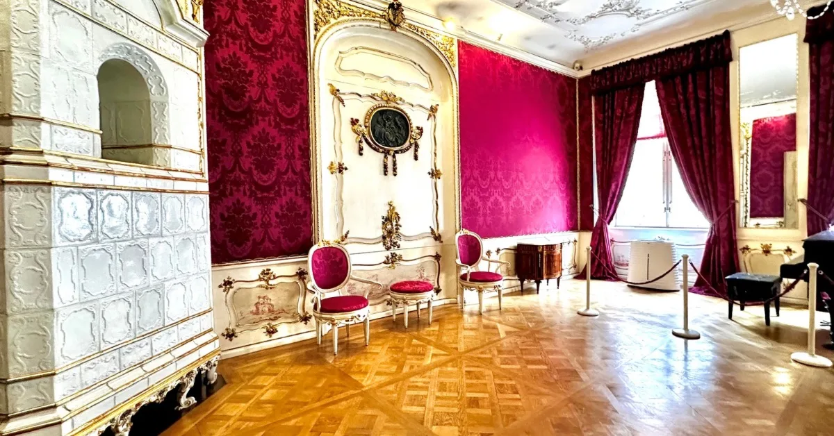 Red wallpapered room, with white tiled fireplace and brown parquet flooring. Red Velvet curtains and red upholstered chairs.