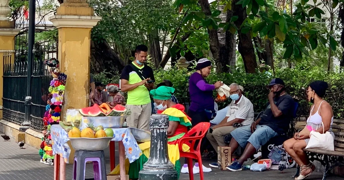 People gathered under trees for shade. Merchants selling local fruits.