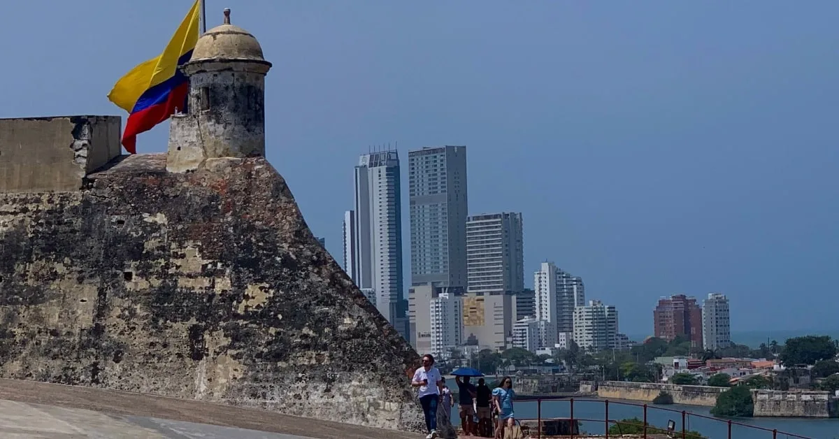 Stone walls of the old Fortress with a Colombia flag flying atop. Across the bay are modern skyscrapers.