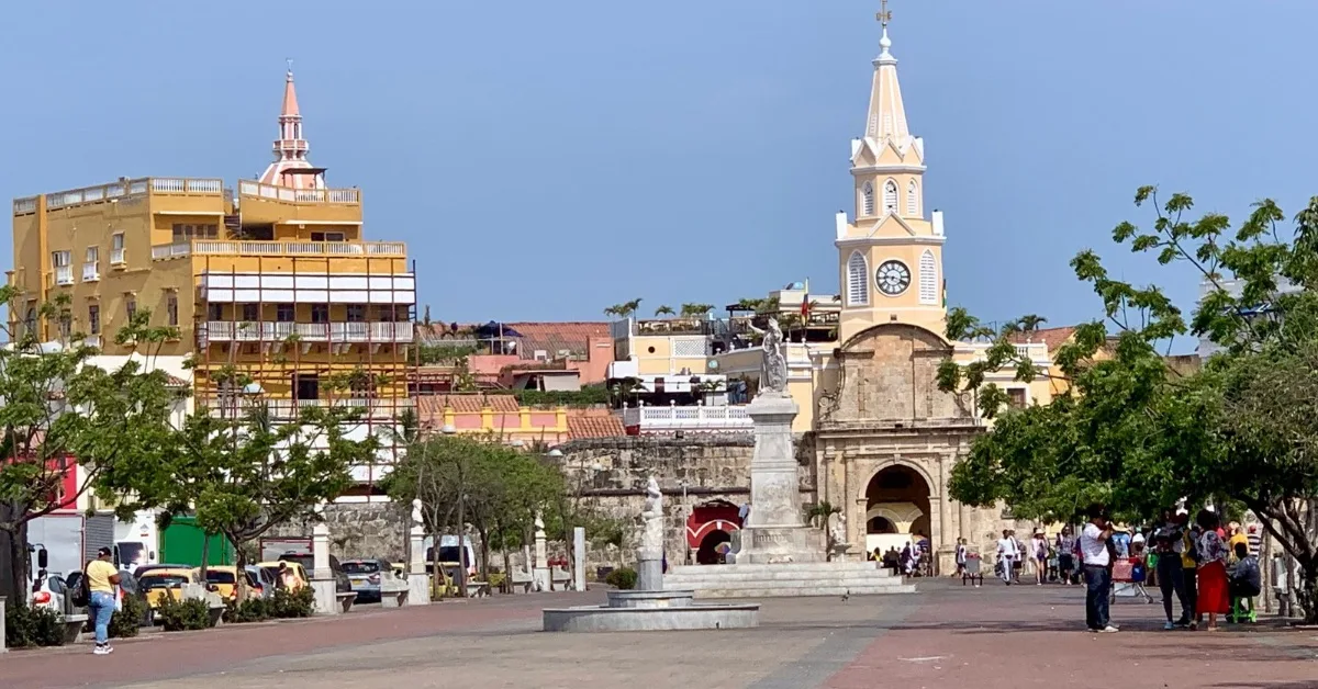 A square between Old City and other parts of Cartagena with people shading from the sun under trees. A clock tower and arch entrance connects the Old Town.