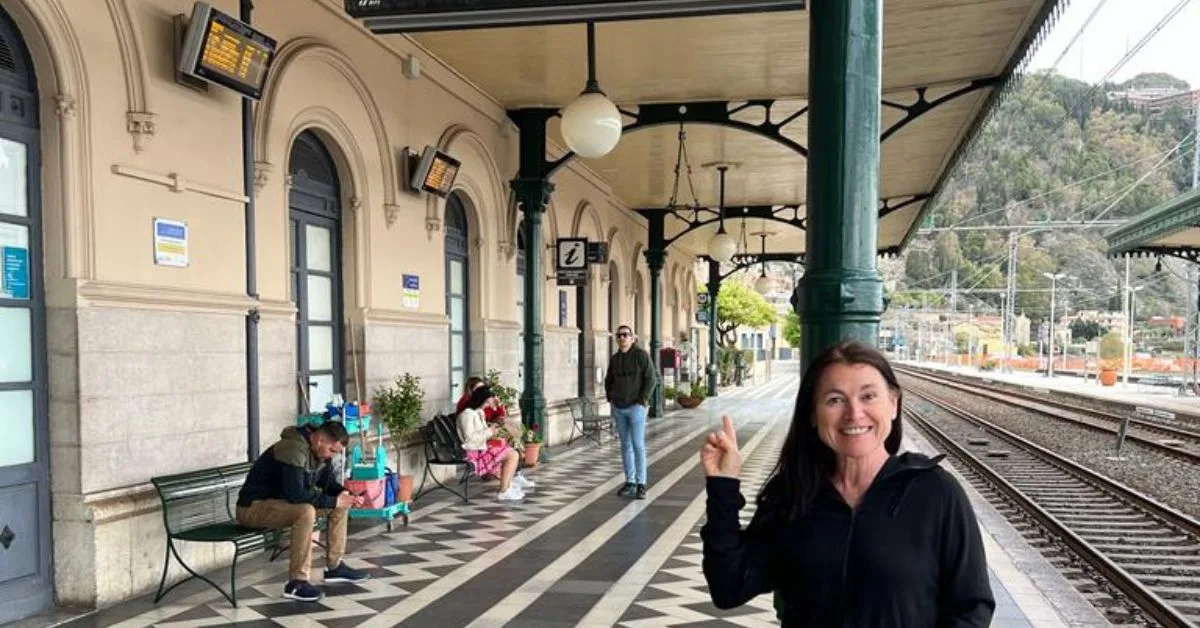 Woman smiling at a train station waiting for her train. Few other people sit on benches in the background.
