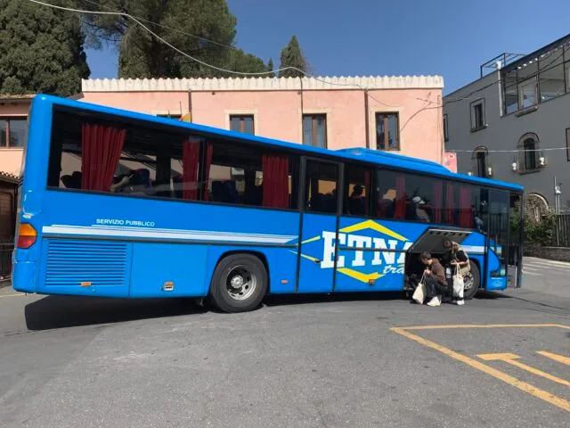 Blue bus running a service to Mount Etna. Two passengers are collecting luggage from the bus storage area.