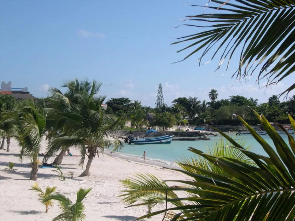 An almost empty white sandy beach with one person walking at the waters edge.