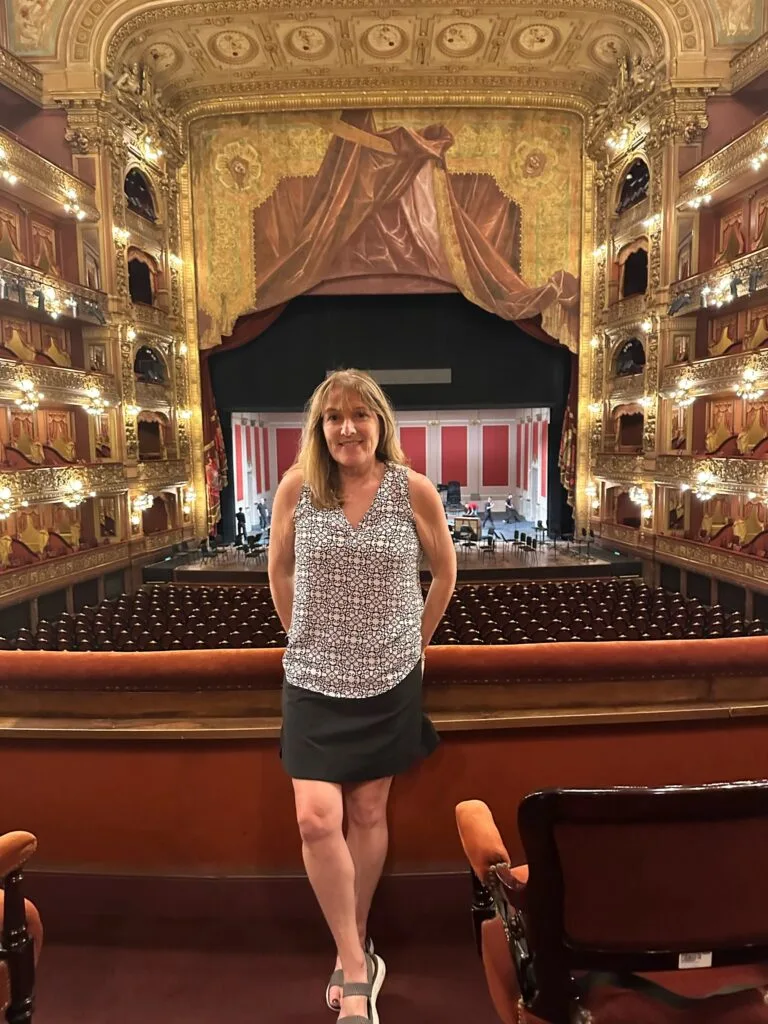 Teatro Colon. Woman standing in the upper seating area and the main stage is behind her. Gold and red colors adorn the theatre.