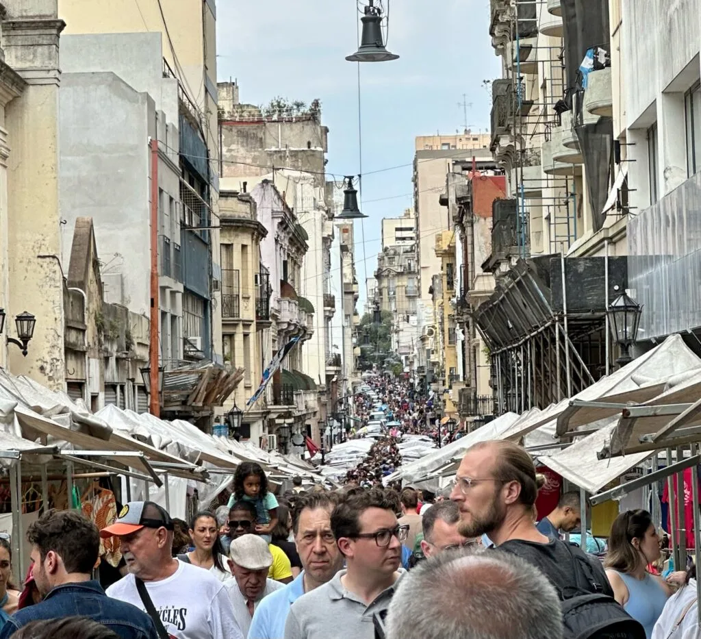 This is a street full of market stalls - more than 600. There are many people enjoying shopping. The market is in old, narrow street with multi-story buildings that have wrought-iron balconies.