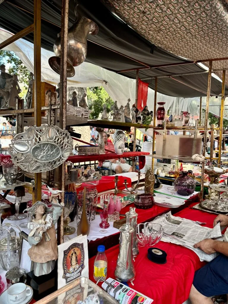 An Antique Stall in San Telmo selling old household items such as figurines, candlesticks, ornamental glass and trays. The items are displayed on 3 shelves.