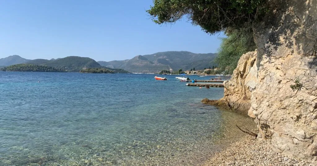 Quiet shingle beach next to calm waters with hills and few boats in the background