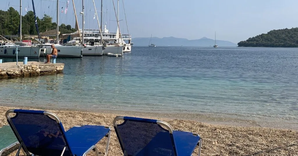 2 blue empty beach chairs sit on beach looking out to calm sea. Peaceful scene with boats in the background