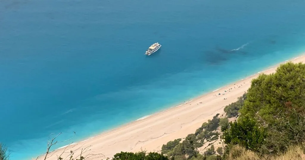 Empty remote beach looking down from road above with one large boat in the sea which is turquoise blue