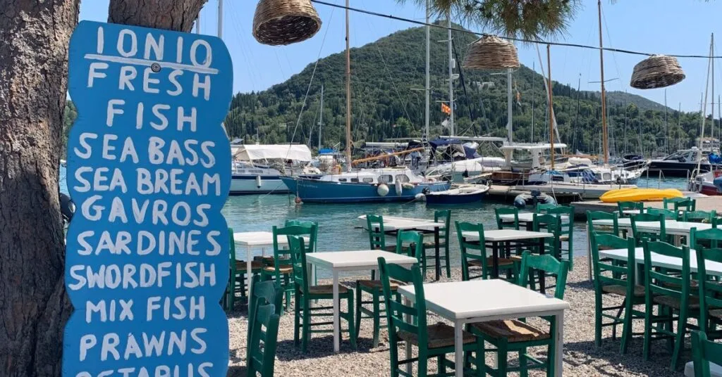 Tables and chairs sit along side harbour for dining and large blue menu is attached to tree showing fish dish choices,