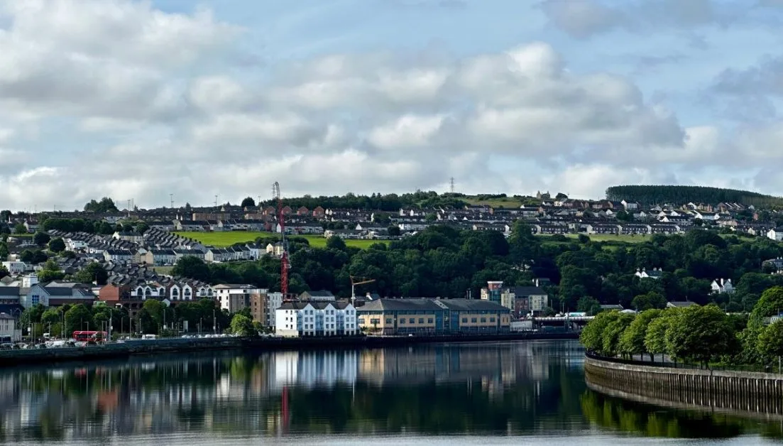 Derry city view from the river with buildings lining one side and hill behind. This is a walled city in Northern Ireland.