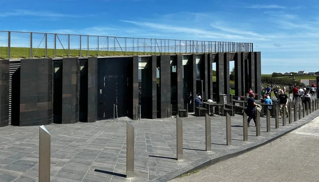 People entering the Visitors Center at Giants Causeway, a steel vertical designed building built into the hillside, with a grass roof.