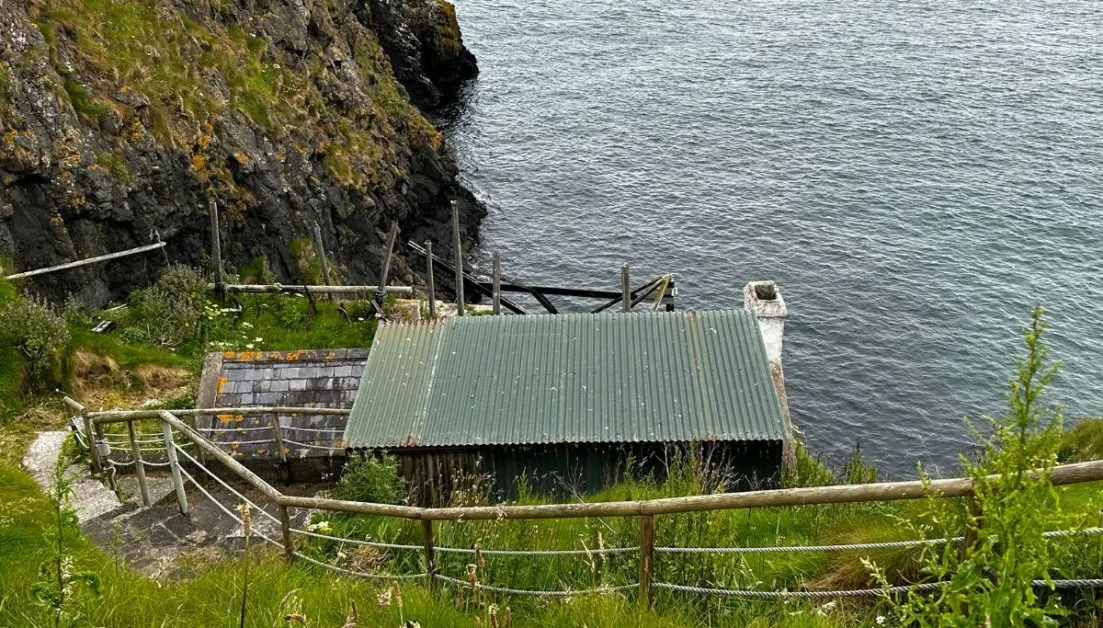 An abandoned fishing cottage built of stone with metal roof built into the rocks above the waters off Northern Ireland.