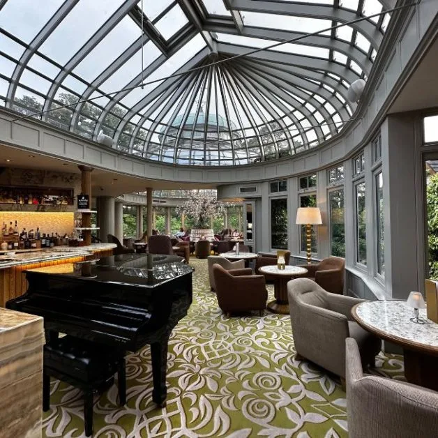 A circular convervatory with glass roof and panorama window set up for guests to enjoy a drink. A piano is in the center of the room.