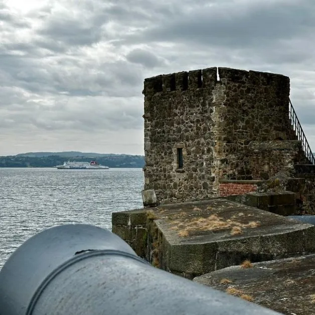 A large canon exhibit on external stone walls of a coastal castle points out to sea. A modern ferry sails past en route to Belfast.