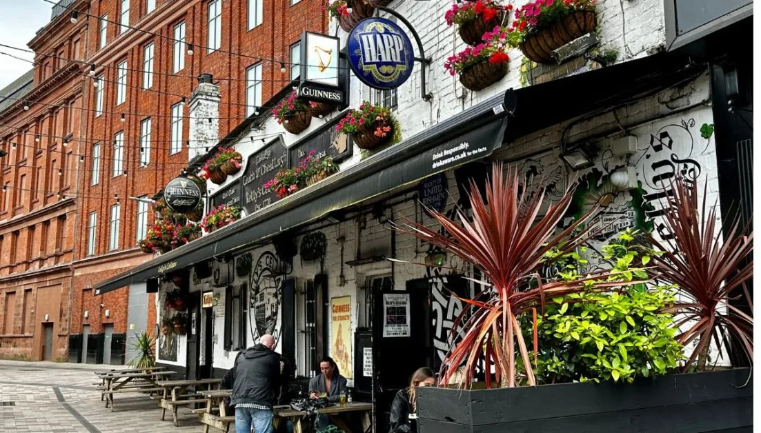 Wooden tables set outside a traditional pub in Belfast. Signs for Guinness hang off the front of the building along with multi-colored flower baskets.