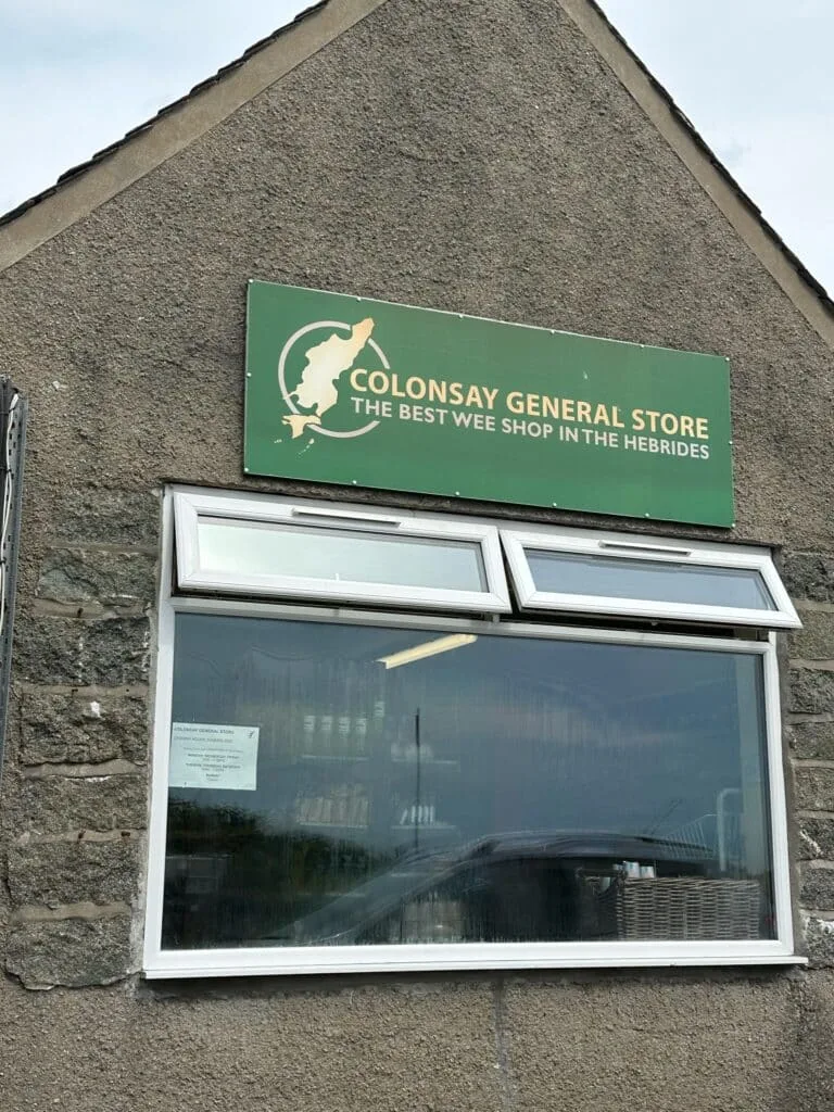A grey building with a large window advertising this is island grocery store and post office on Colonsay Island, Scotland.