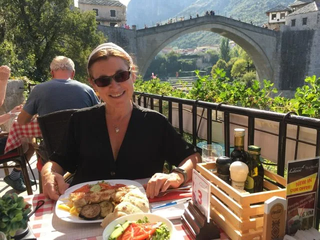 Women eating at an outdoor cafe and behind her is the Stari Most bridge in Mostar .