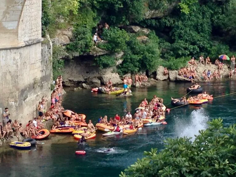 Families on floats on a river, and sitting under a bridge structure in Mostar.