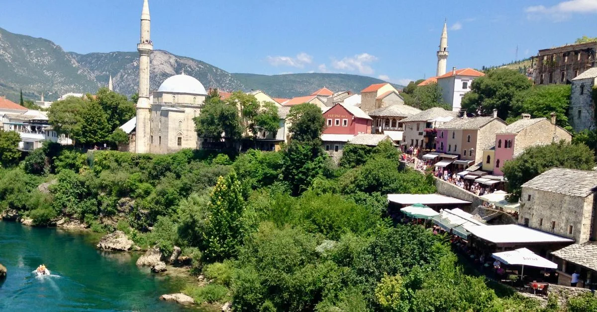 A Sarajevo mosque sits near to a river and leads down to a medieval cobbled street where tourists are walking.