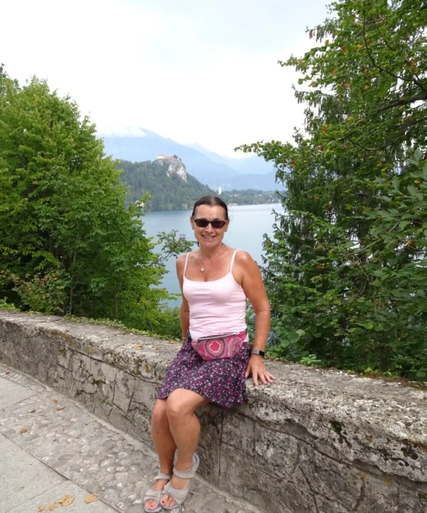 Woman sitting on a stone wall at Lake Bled. Trees surround the lake and mountains are visible in the distance.