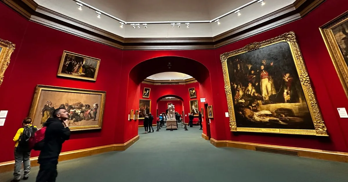 Large national painting hang on a rich red wall in the national gallery of Scotland. People are admiring them.