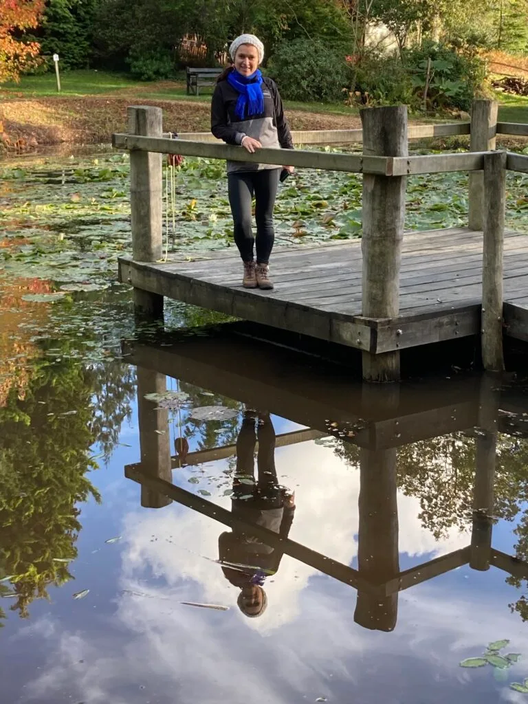 A woman stands next to a pond reflecting in the stillness of the waters.