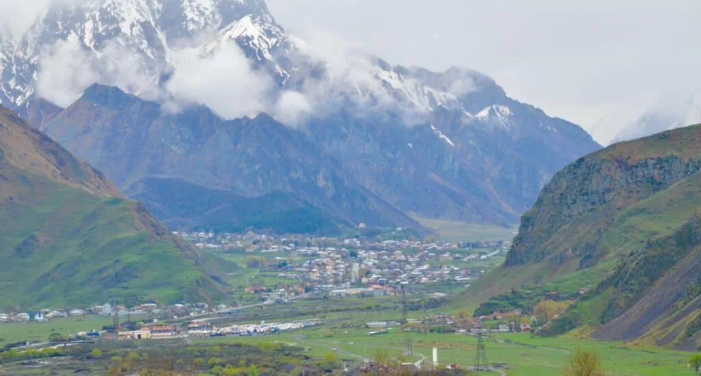 View of Kazbek Mountain with snow capped peaks and a town in the valley below.