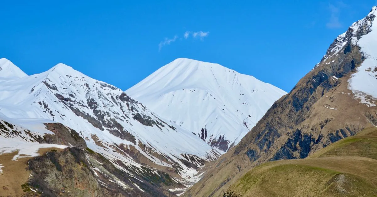 View of snow capped peaks and rock faces at lower levels, some with grazing grass on steep slopes.