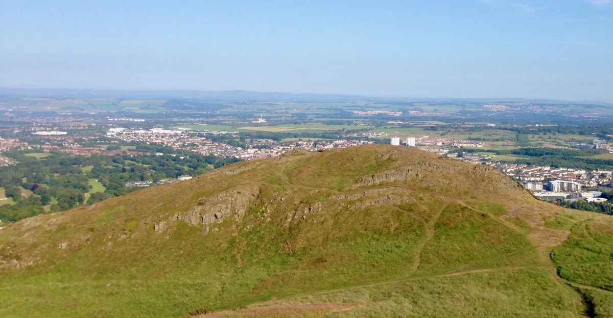 View of Edinburgh from Arthur's Seat with grassy and rock exposure hill tops and behind them city scenes.