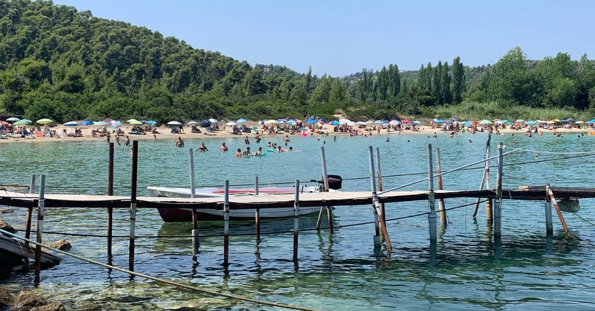 Wooden bridge on stilts with boat behind it on the calm water. People are in the water and strip of beach can be seen in the distance