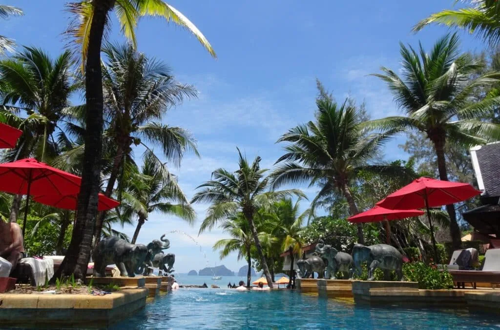 A swimming pool with sun loungers and red parasol shade. Palm trees adorn the area and it looks out to 0pen waters.