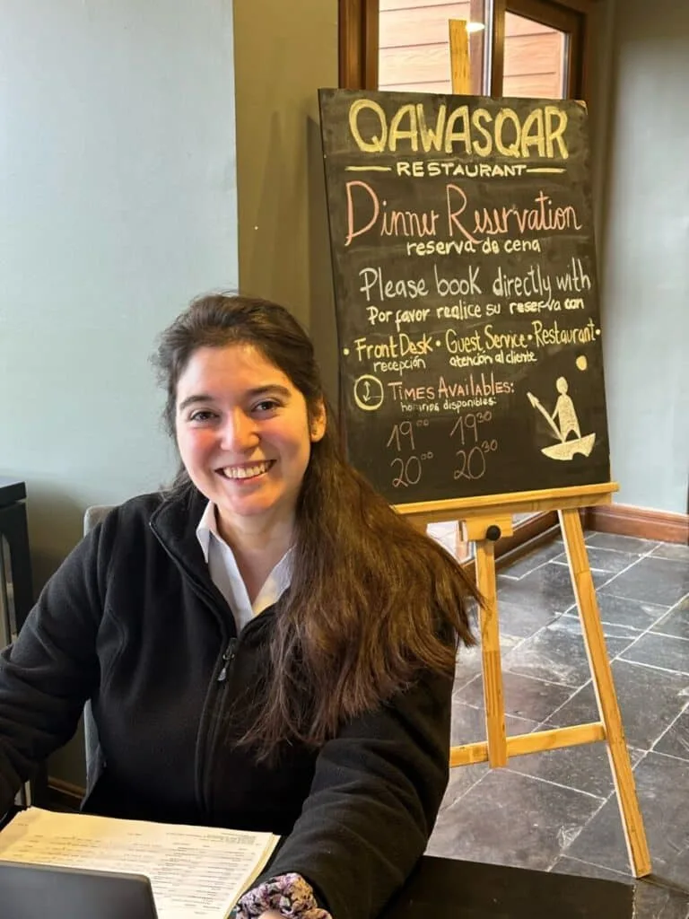 Woman is at a reception desk in a hotel with a blackboard behind her showing information for staff.