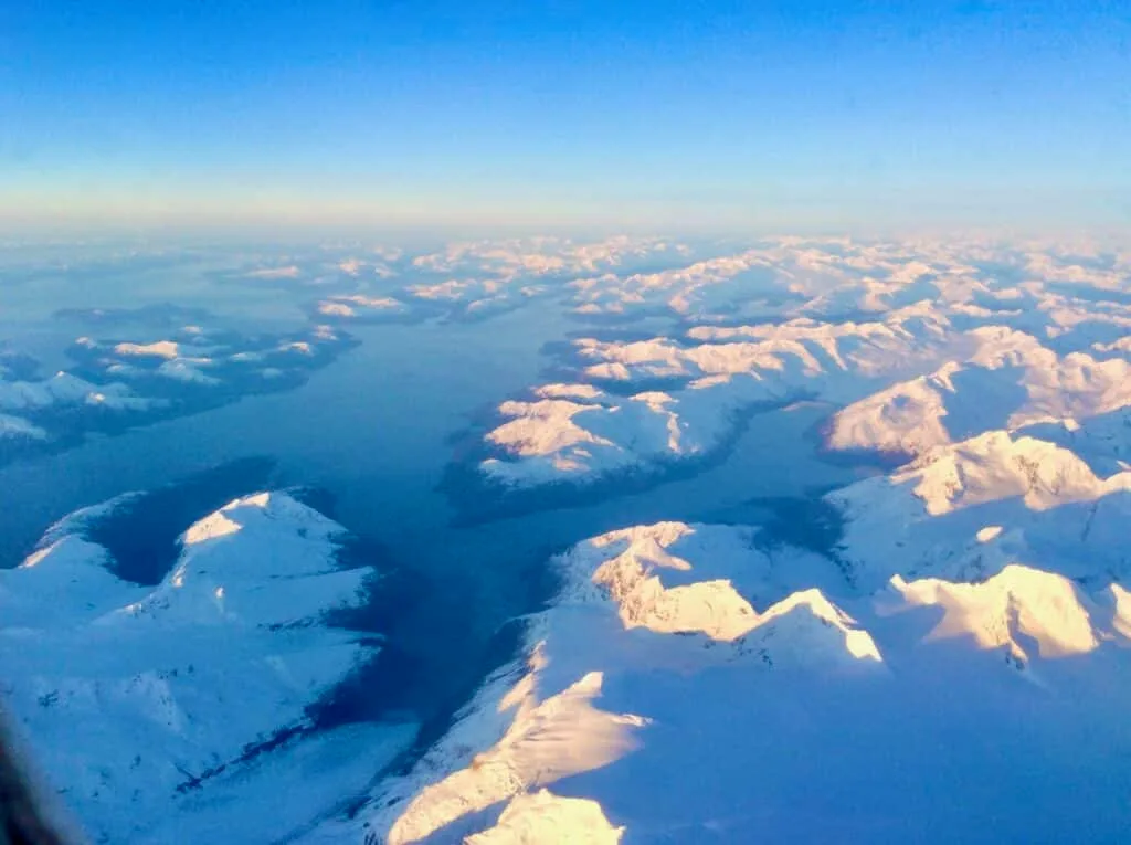 Aerial view of large inlets in Alaska covered in snow and glaciers. Blue waters are reflected where ocean meets land.