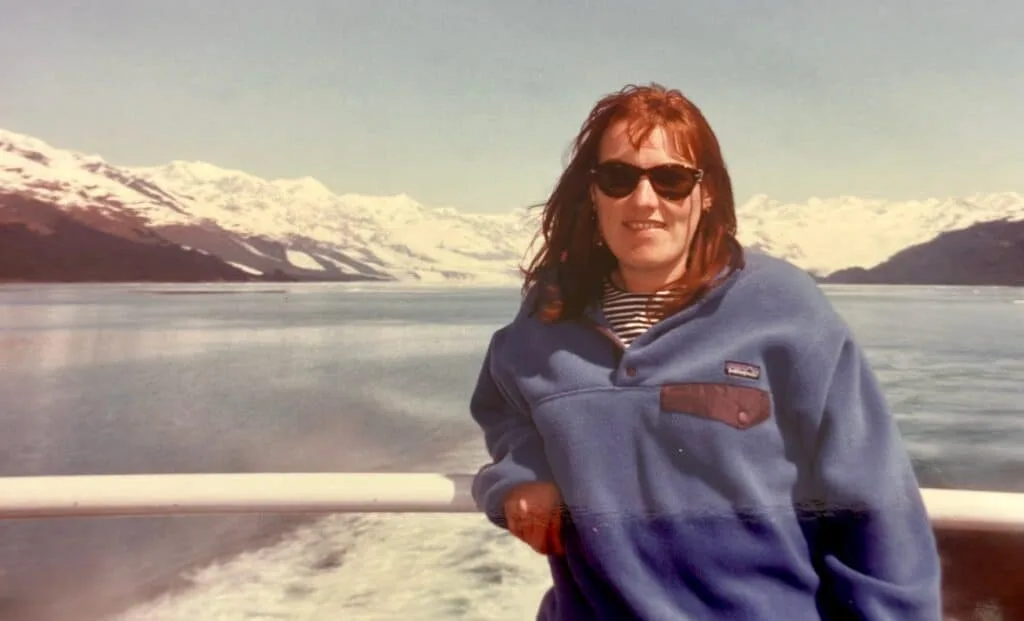 Woman is standing on the deck of a tourist boat in Alaska. Mountains and glaciers are visible in the distance.