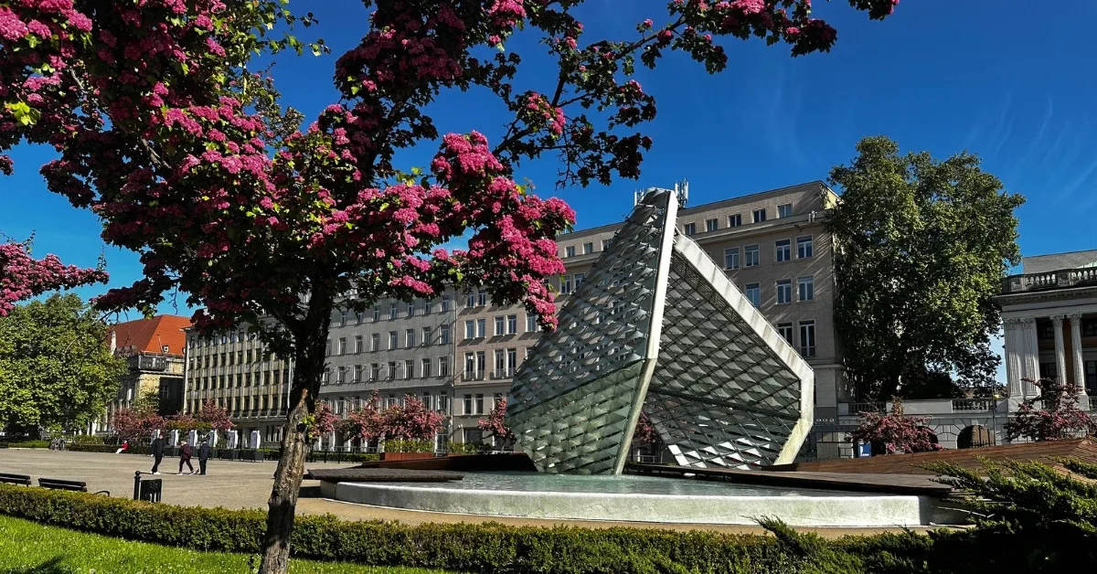 A large metal structure in a fountain is a memorial within Freedom Square in Poznan. Pink blossom is on the trees that line the square.