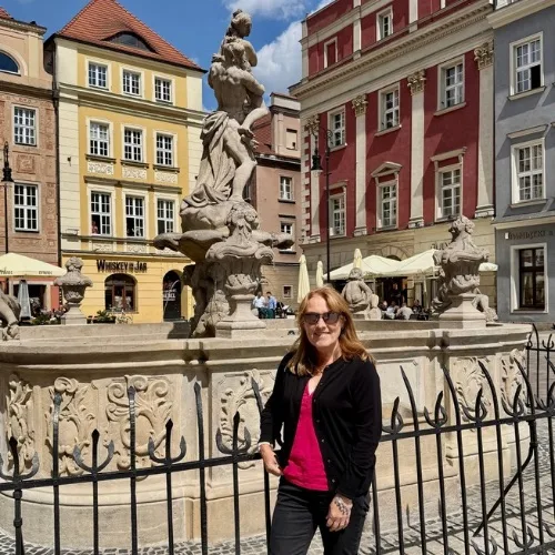 Woman standing in front of the main fountain in the Old Market Square Poznan. Behind her are classic 3-4 story baroque buildings painted in different colours including yellow and red.