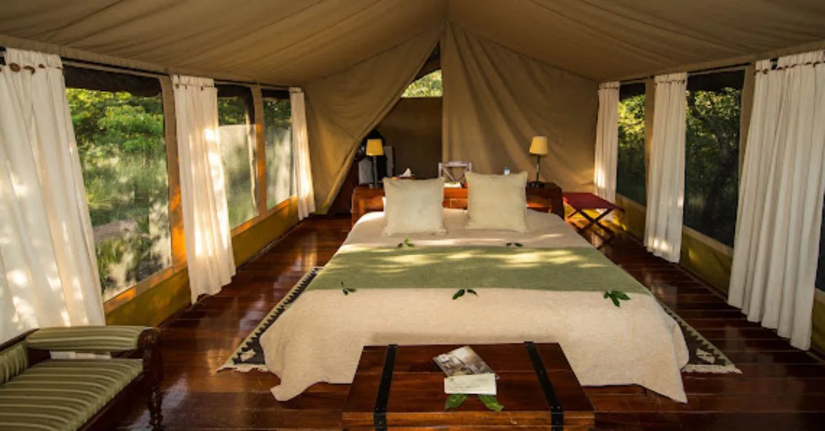 Interior of a tent at Karen Blixen Camp shows bed, seating area and curtains. The floor is wooden with rugs either side of the bed.