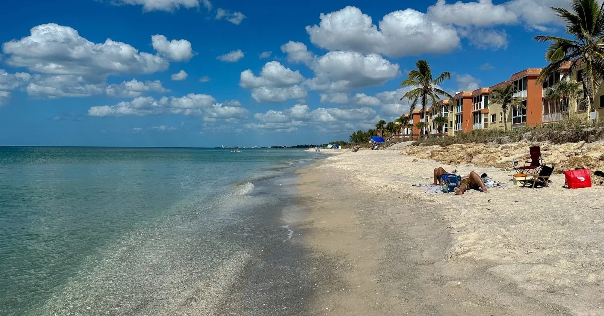 Sandy beach with a resort building and 2 people on deck chairs.