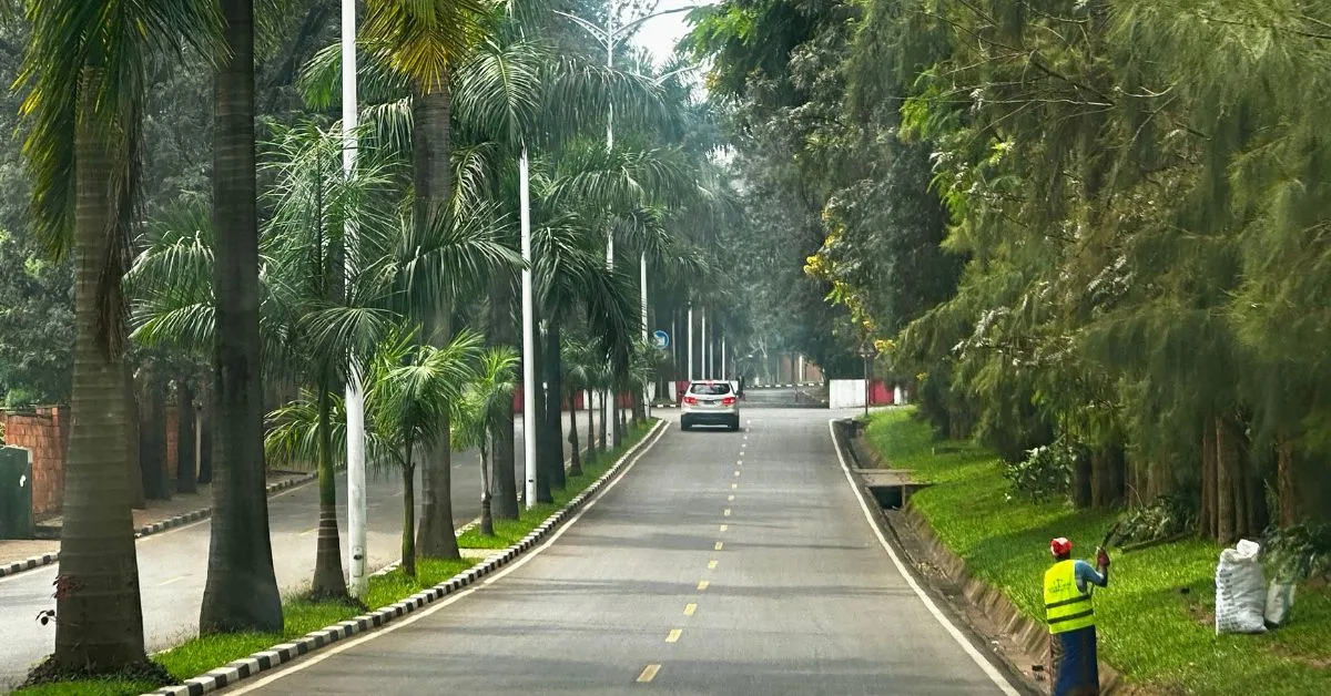 A Main Street in Kigali is lined with trees and lawns. A person is sweeping up leaves and other debris.