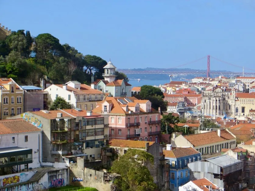 Views of Lisbon with colourful red tiled homes, churches and other buildings. The view is also of the river Tagus and a suspension bridge.
