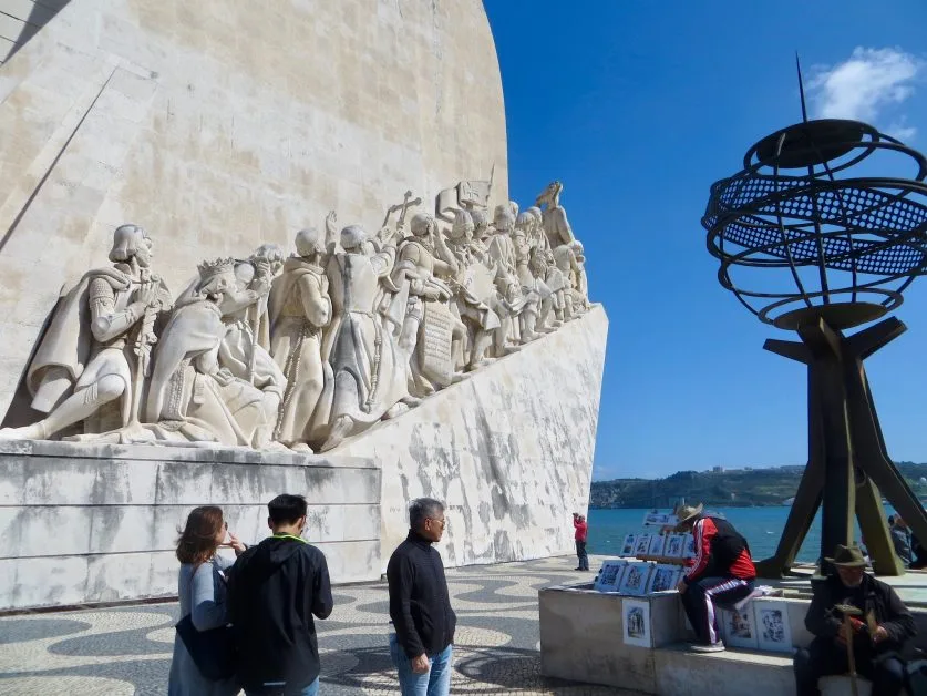 A large monument to celebrate where many Portuguese explorers left the country centuries ago. There is an artist selling his art work near the base of the monument and several tourists walking around.